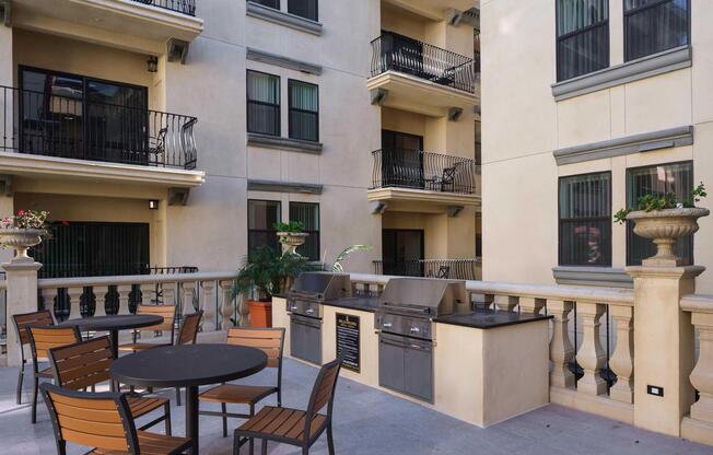 Outdoor lounge area featuring a large table with chairs, two barbecue grills, and decorative potted plants. The backdrop includes balconies of a multi-story building with wrought iron railings, creating a welcoming space for residents to gather and socialize.