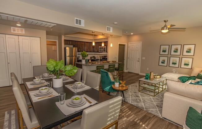 A modern living space featuring a dining area with a large table set for six, green chairs, and decorative plants. The open kitchen includes stainless steel appliances and dark cabinetry. The living room showcases a comfortable sofa, coffee table, and wall art, all in a stylish and inviting color scheme.