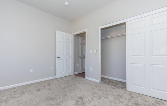 A well-lit room with light gray walls and carpeted flooring. There are two closed white doors on the left leading to a closet and an entrance. A third door is visible in the background, suggesting access to another area. The space appears clean and spacious, suitable for various uses.