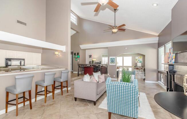 Spacious living area featuring a modern open layout, tile flooring, and tall ceilings. Two gray barstools at a countertop area, a light gray sofa with colorful throw pillows, and a stylish blue checkered chair. Large windows allow for natural light, creating an inviting atmosphere.