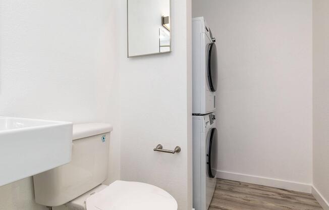 A small laundry room featuring a stacked washer and dryer, a white toilet, and a small sink. The room has light-colored walls and a simple, modern design. A mirror is mounted on the wall beside the toilet, and the flooring is a light wood finish.