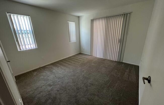 A vacant room featuring light-colored walls and two narrow windows with white blinds. The floor is covered with gray carpet, and there is a sliding glass door on one side leading to an outdoor area. The room appears bright and spacious but unfurnished.