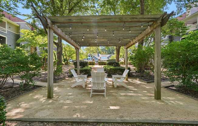 A wooden pergola with white chairs is in the middle of a sandy area.