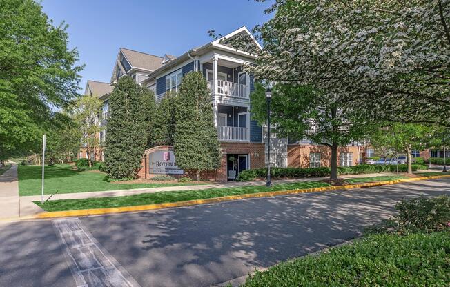 A view of a modern apartment building surrounded by greenery, featuring well-maintained landscaping and a sign indicating "Rothbell." The image captures a peaceful residential area with trees lining the street and a clear blue sky overhead.