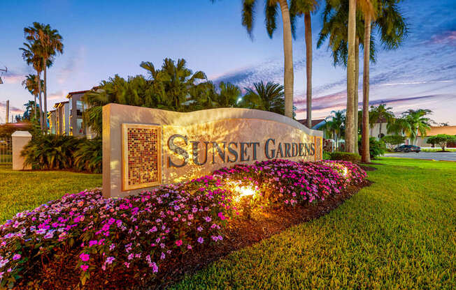 Sunset Gardens sign with flowers in front and palm trees in the background.