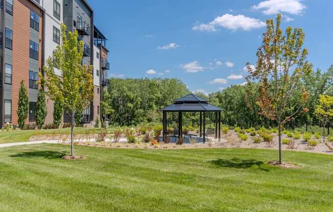 A gazebo sits in the middle of a grassy area with trees and apartment buildings in the background.
