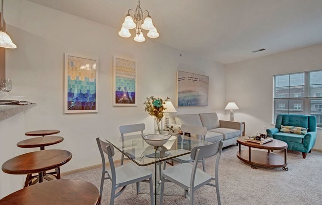 A bright dining and living room here at Swan Creek with beige carpet, gray sofa, and teal accent chair. Glass dining table and open layout create a comfortable and inviting space.