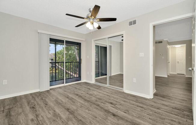 Spacious interior of a modern apartment, featuring a ceiling fan, large mirrored closet doors, and sliding glass doors leading to a balcony. Light-colored walls and laminate flooring create a clean, inviting atmosphere. A hallway is visible in the background, leading to additional rooms.