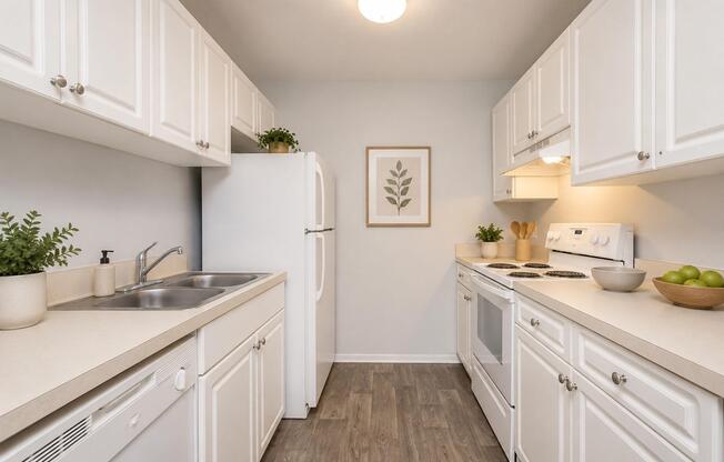 A bright and modern kitchen featuring white cabinets, a double sink, and a white refrigerator. The countertops are light-colored with a bowl of green apples. An oven and stovetop are present, along with decorative plants and a framed leaf print on the wall, creating a clean and inviting atmosphere.