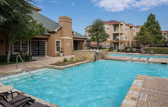 A large outdoor swimming pool with a stone border and a small patio area with chairs and a table.