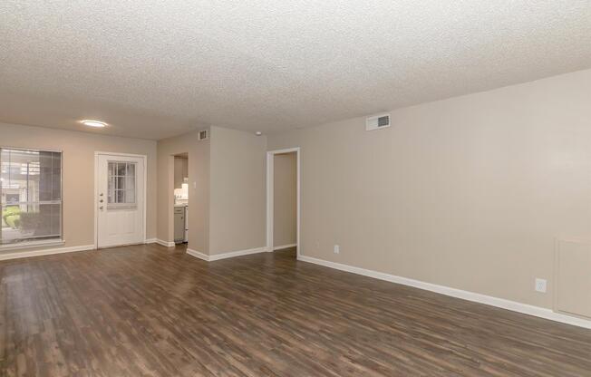 Spacious living area with light-colored walls and a textured ceiling. The room features laminate flooring, a front door with windows, and a doorway leading to a kitchen. A hallway to another room is visible, providing an airy and bright atmosphere.