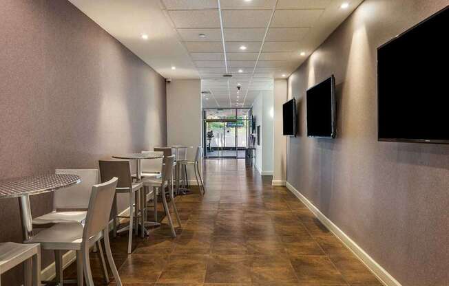 A long hallway with chairs and tables on the left and two TVs on the wall.