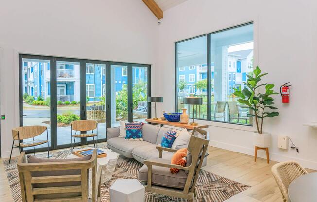 A modern living room featuring a gray sofa with colorful throw pillows, a patterned rug, and a coffee table. Large windows provide natural light and a view of greenery outside. There are two stylish chairs, a potted plant, and a fire extinguisher on the wall, creating a cozy and inviting atmosphere.