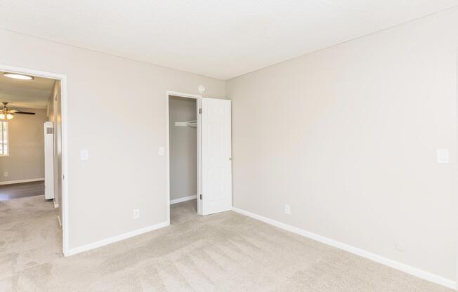 A well-lit, empty room with beige walls and light beige carpeting. It features a closed door leading to a closet on one side and an open doorway leading to another room on the opposite side. The ceiling has a textured finish, and there is a small wall outlet visible. Natural light enters from a window out of view.