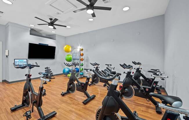 A room with a white wall and a ceiling fan with a row of stationary bikes in front of it.