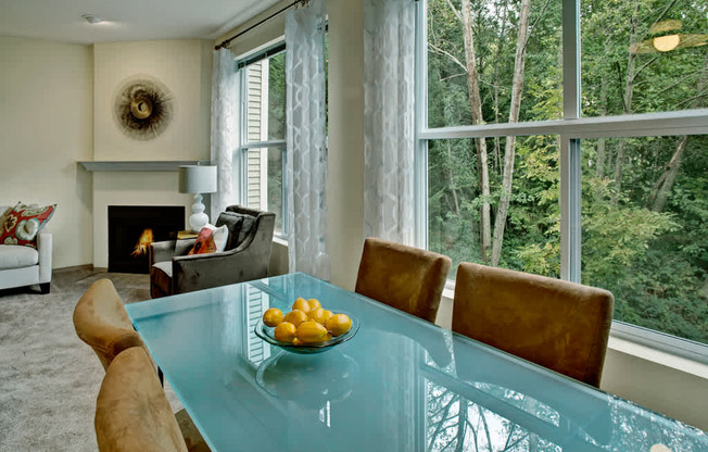 A light-filled dining area here at Portsmith featuring a glass-top dining table with upholstered chairs set beside expansive windows with sheer patterned curtains, showcasing leafy wooded views, and opening into a cozy living space with soft carpet flooring, an accent chair, decorative wall art, and a fireplace that adds warmth and visual interest.