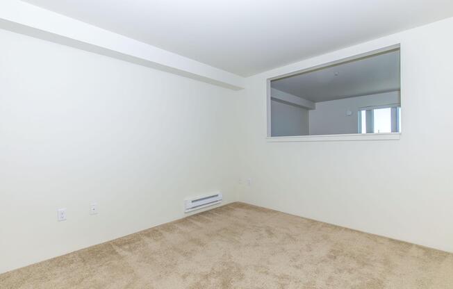 A sparsely furnished room with light beige carpet and white walls. There is a large window with a mirror above it, allowing natural light to enter. The space appears empty and clean, suitable for personalization or decoration.
