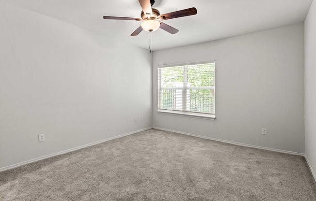 A bright, empty bedroom featuring light gray walls and a ceiling fan. The room has soft beige carpet flooring and a large window with natural light coming through, providing a view of greenery outside. The space is uncluttered, creating a fresh and airy atmosphere.
