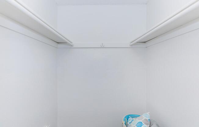 A small, empty closet with plain white walls and two hanging rods. In the corner, there is a mesh bag containing items, and the floor is bare. The overall appearance is clean and uncluttered, suggesting minimal storage use.