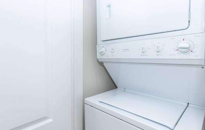 A stacked laundry unit featuring a washing machine on the bottom and a dryer on top, with control knobs and settings visible. The unit is positioned against a light-colored wall, with a door slightly visible nearby.