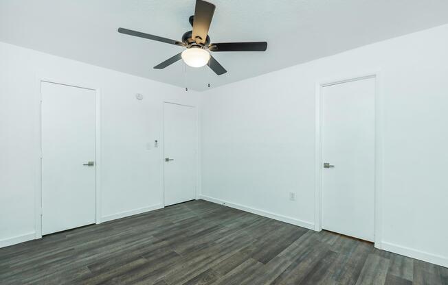 Empty room with light gray walls and a ceiling fan. Two white doors lead to adjacent spaces. The floor is a dark laminate, providing contrast to the walls. The room is well-lit and has a clean, minimalistic aesthetic, suitable for various uses.