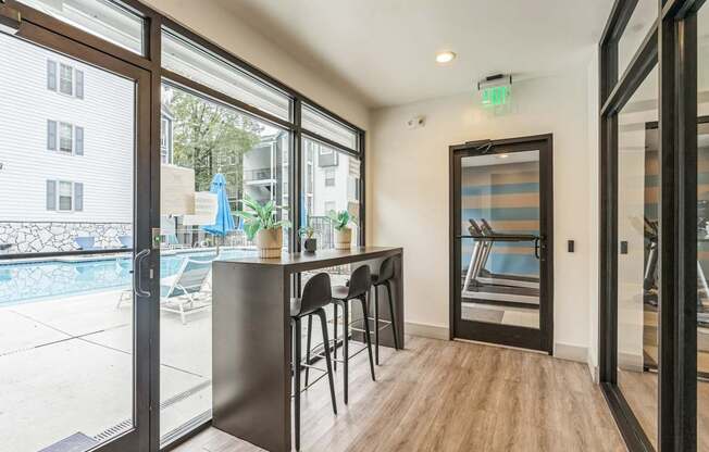 A room with a bar and chairs is shown with a view of a pool and buildings outside.