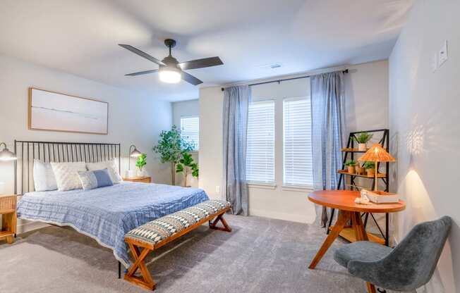 Two-Bedroom Apartments in Raymore, MO - The Venue Townhomes - Bedroom with Carpeted Flooring, Bed, Footboard Bench, Wooden Nightstands with Lamps, House Plants, Circular Wooden Table, Grey Office Chair, Ceiling Fan, Display Shelf, and Large Windows.