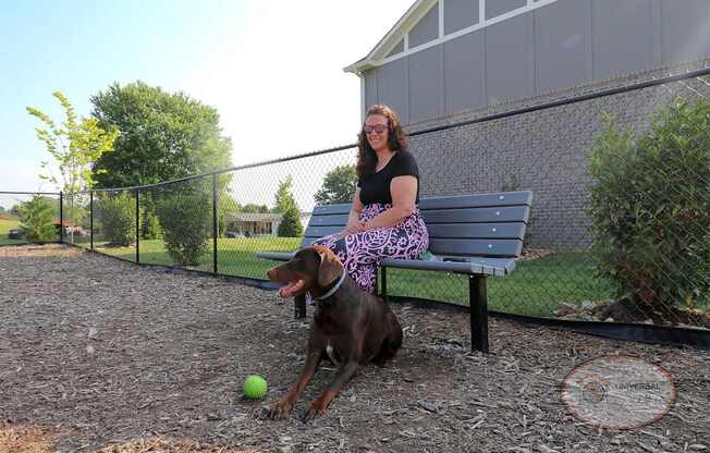 A woman sits on a bench with a her dog inside a fenced in dog park.
