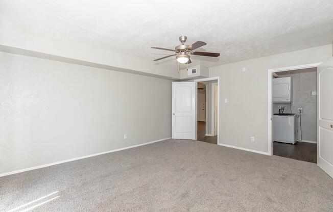 A spacious, well-lit room with light gray walls and plush carpeting. Features a ceiling fan and an open door leading to a laundry area. The room has a neutral color palette, making it feel warm and inviting. Natural light streams in, enhancing the overall ambiance.