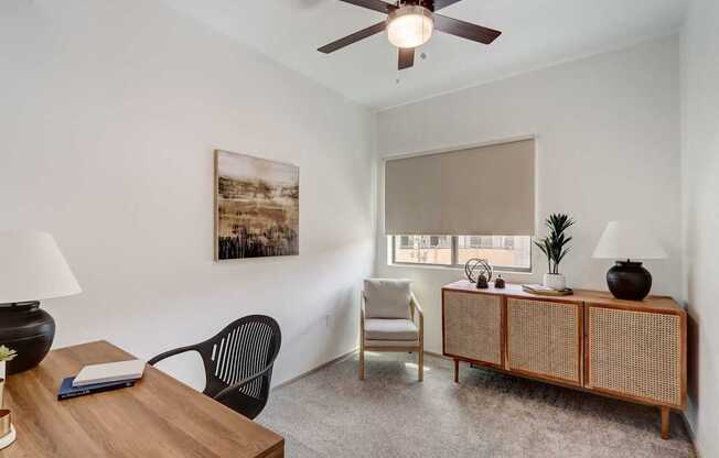 A room with a ceiling fan, a chair, a table with a book and a lamp, and a painting on the wall.