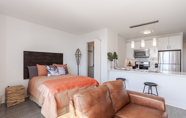 A bedroom with a bed, a couch, and a kitchen area in the background.