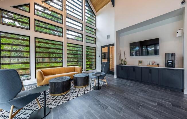 A community lounge with a sofa, two chairs, coffee tables, and large windows near a resident coffee station with a TV.