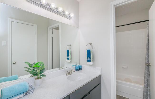 A modern bathroom featuring a double vanity with a large mirror, a stylish light fixture, and a white countertop. There are neatly arranged towels and a small potted plant on the counter. A shower curtain indicates a bathtub/shower area is on the right, and a door leads to another room. The decor is clean and minimalistic.