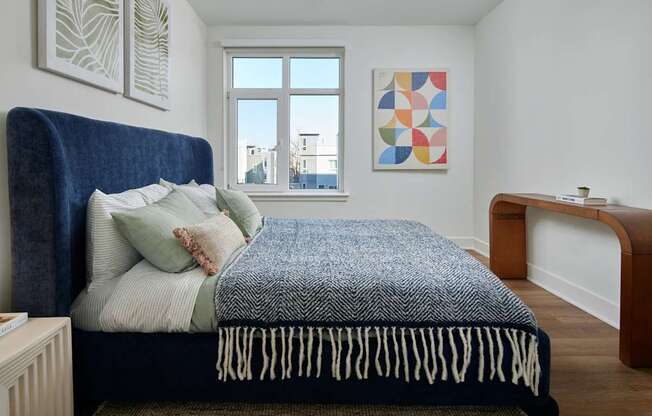 A bed with a blue headboard and a white and black blanket is in the foreground of a room with a window and a painting on the wall.