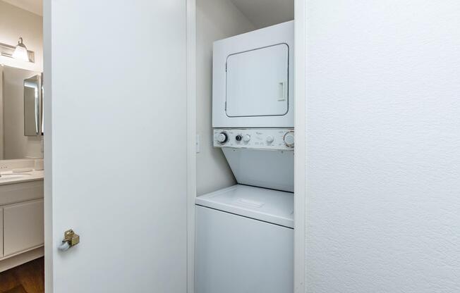 A compact laundry area featuring a stacked washer and dryer unit, located in a small room with white walls and a door partially open. In the background, a glimpse of a bathroom with a mirror and light fixture can be seen. The floor is wooden, adding warmth to the space.