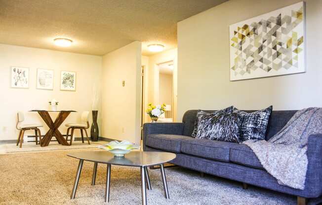 Carpeted living room with hallway that leads to bedrooms. Staged with grey couch, coffee table and art.at The Lakes Apartments, Washington, 98837