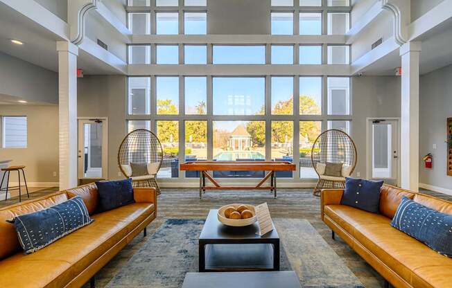 A spacious resident lounge here at Retreat at McAlpine Creek featuring leather sofas with accent pillows, a central coffee table, and a shuffleboard table positioned near floor-to-ceiling windows. Hanging lounge chairs add visual interest, while abundant natural light and views of the pool area create an open, inviting space for relaxing or socializing.