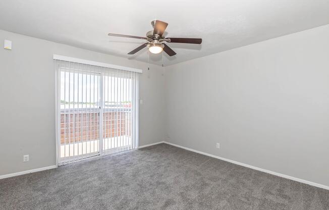 A spacious, empty living room featuring gray carpet flooring, a ceiling fan with wooden blades, and sliding glass doors leading to a balcony. The walls are painted light gray, and there is natural light coming through the blinds. The room has a clean, modern aesthetic.