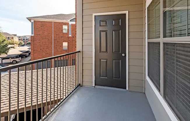 A balcony here at Landing at Mansfield with gray floor, black railing, tan siding, dark door, and a window with white blinds overlooking nearby buildings and parking.