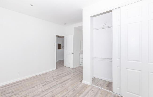 A bright, minimalistic room with light-colored walls and wooden flooring. A closet with sliding white doors is on the right, partially open, revealing shelves inside. A doorway in the background leads to another area, creating an open and spacious feel.