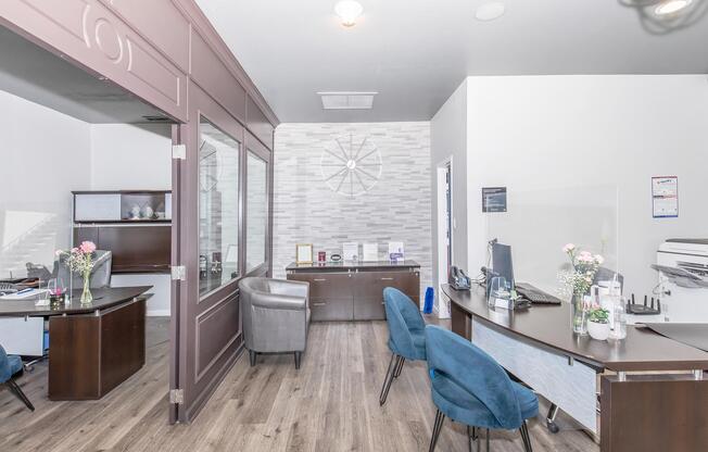 A modern office interior featuring a reception area with a desk, chairs, and decorative elements. The space is well-lit with gray and white walls, wooden flooring, and a large wall clock. Flowers in vases add a touch of warmth, creating a welcoming and professional atmosphere.