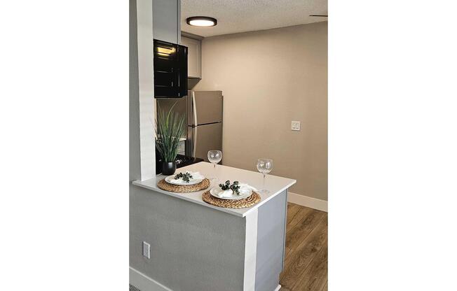 A modern kitchen nook featuring a countertop with two decorative plates, each topped with a small plant, and two wine glasses. In the background, there is a refrigerator and a neutral-colored wall. The floor has wood-like laminate, adding to the contemporary feel of the space.