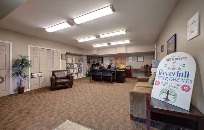 A sign in the foreground of a room with a carpeted floor and a few chairs.