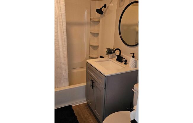 A modern bathroom featuring a gray vanity with a white countertop, a round mirror, a black faucet, and a small plant. The bathtub has a white shower curtain, and there are built-in shelves visible. The floor is covered with a black mat, and a toilet is positioned to the right.