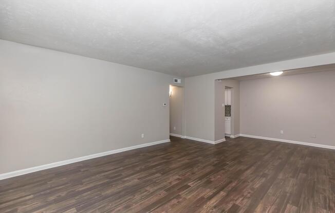 A spacious, empty living area with light-colored walls and a dark wood laminate floor. There is a doorway on the left leading to another room, and a small opening on the right suggesting access to a kitchen or dining area. The room is well-lit with a ceiling light fixture.