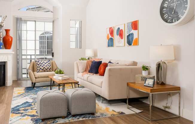 Apartments for Rent in Miracle Mile Los Angeles, CA - The Preston - Living Room with Plank Wood Style Flooring, Couch, Armchair, Ottomans, Side Tables with Lamps, Coffee Table, Fireplace, Framed Art, Wall Clock, Window, and Sliding Glass Door with Access to Patio/Balcony