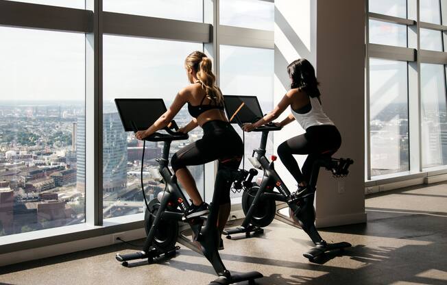 Two women are exercising on stationary bikes in a bright room with a city view.