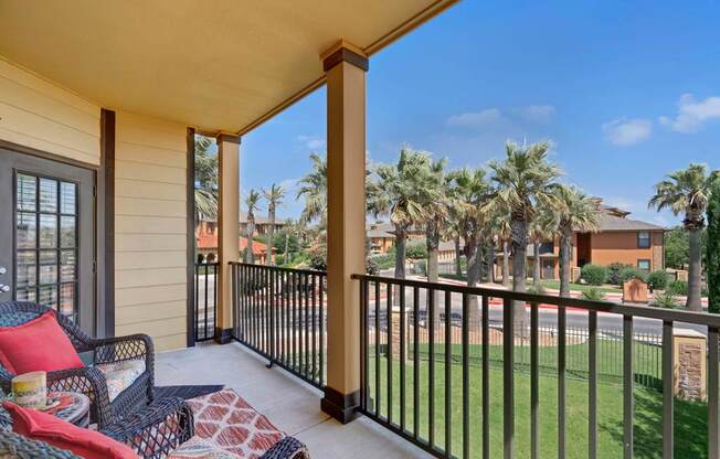 A covered private balcony here at Tuscany at Faudree with outdoor seating arranged on a concrete floor. A railing overlooks landscaped grounds with palm trees and nearby community buildings, while the shaded layout offers a comfortable space to relax and enjoy fresh air and neighborhood views.
