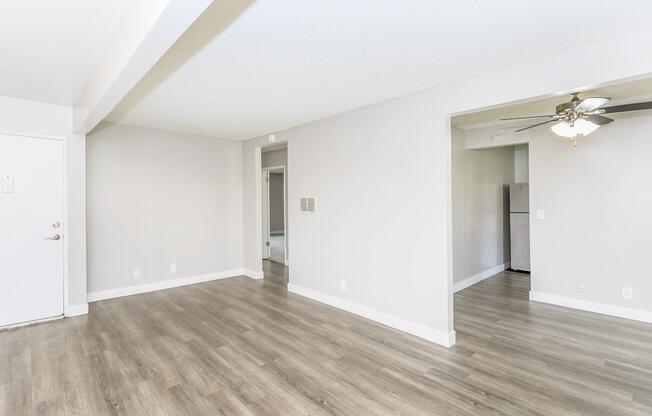 A spacious, light-filled living area featuring light-colored walls and wood-like flooring. A doorway leads to another room, and a ceiling fan is visible in the corner. The overall ambiance is modern and minimalistic, with ample natural light.