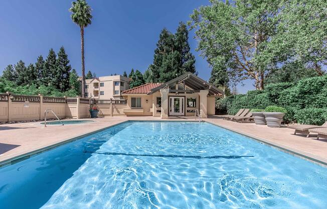 A clear blue swimming pool surrounded by palm trees and lush greenery. In the background, a small building with a tiled roof and large windows overlooks the pool area, which features lounge chairs and a serene atmosphere under a bright blue sky.